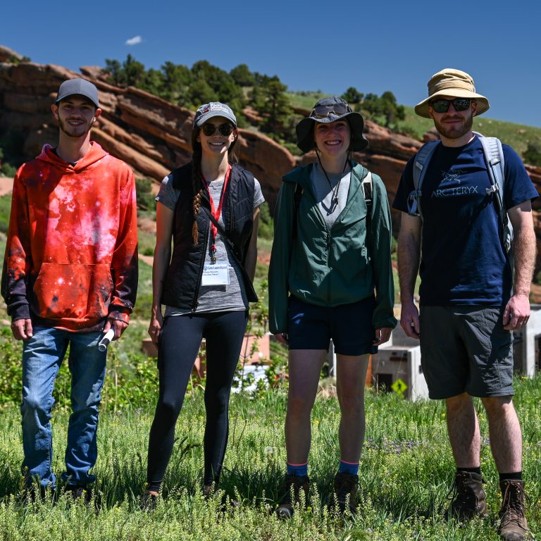 The four 2021 Geo-Launchpad interns standing in a line in front of a large outcrop at Red Rocks Park in Morrison, CO.