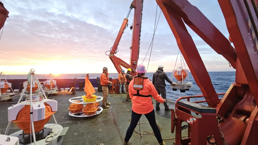 Crew on the deck of a ship use a crane to lower round, orange instruments into the water.